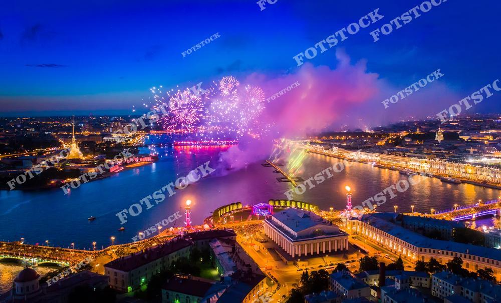 На фотографии изображён праздничный фейерверк над Невой в Санкт-Петербурге. На переднем плане видны Ростральные колонны и здание Биржи на Стрелке Васильевского острова. Вдали виднеется Петропавловская крепость, освещённая огнями. Город погружён в атмосферу праздника и сияет в ночных огнях.